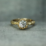 A yellow gold ring with a lab-grown diamond in a decorative bezel setting, resting on a textured gray surface.