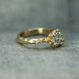 A gold ring with a textured, organic-shaped band and a lab-grown diamond in a bubbly ocean setting, resting on a gray stone surface.
