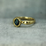 A gold ring with a textured, organic-shaped band and a bezel oval-set dark blue sapphire, resting on a gray textured surface.