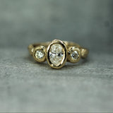 A yellow gold ring featuring a large oval center lab-grown diamond flanked by two smaller round  lab-grown diamonds, photographed on a textured gray surface.