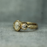 A gold ring with a large lab-grown diamond and a smaller round-cut lab-grown diamond, photographed on a textured grey background.