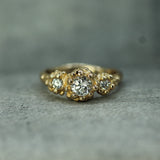 A yellow gold ring featuring a central round lab-grown diamond surrounded by two smaller lab-grown diamond clusters, set against a textured gray background.