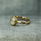 A yellow gold ring with a textured band and a bezel-set oval lab-grown diamond, resting on a gray textured surface.