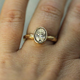 A close-up of a gold ring with an oval lab-grown diamond worn on a person's finger.