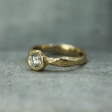 A gold ring with a round-cut lab-grown diamond in a bezel setting, featuring an organic, textured wavy band, photographed on a neutral gray surface.