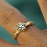 A close-up of a yellow gold ring with a round brilliant-cut lab-grown diamond set in oceany bubbles worn on a finger against a soft teal background.