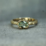 A yellow gold ring featuring a central oval-cut light green sapphire in a bubbly ocean setting, with small accent bubbles along the band, resting on a textured gray surface.