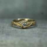 A gold ring with a textured, organic ocean inspired style band and a round clear gemstone in the center, resting on a textured gray surface.