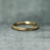 A textured gold band ring with an organic, bark-like surface, resting on a gray stone surface.