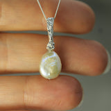A close-up of a silver pendant necklace with a pearl, held between fingers. The silver bail features a small clear stone, and the pearl has a pearlescent cream to light green iridescence.