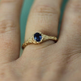 A close-up of a hand wearing a gold ring with a round dark blue sapphire.