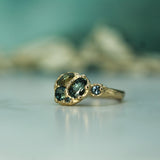 A gold ring featuring multiple sapphires in a textured setting, with a smaller round blue stone on the band, photographed on a light surface with a soft teal background.