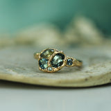 A gold ring with three oval blue-green gemstones, resting on a textured light beige stone surface with a soft teal background.