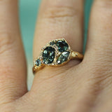 A gold ring with dark green marquise-cut gemstones and small clear accent stones, shown worn on a person's finger.