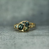 A 14k yellow gold ring featuring multiple sapphires in a textured bezel setting, resting on a textured gray surface.