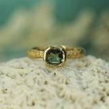 14k yellow gold engagement ring with a teal sapphire on a textured surface with a blurred natural background