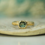Gold engagement ring with a teal sapphire  on a blurred natural background