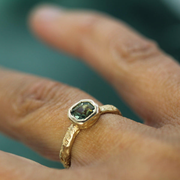 Gold ring with a teal sapphire on a finger against a blurred background