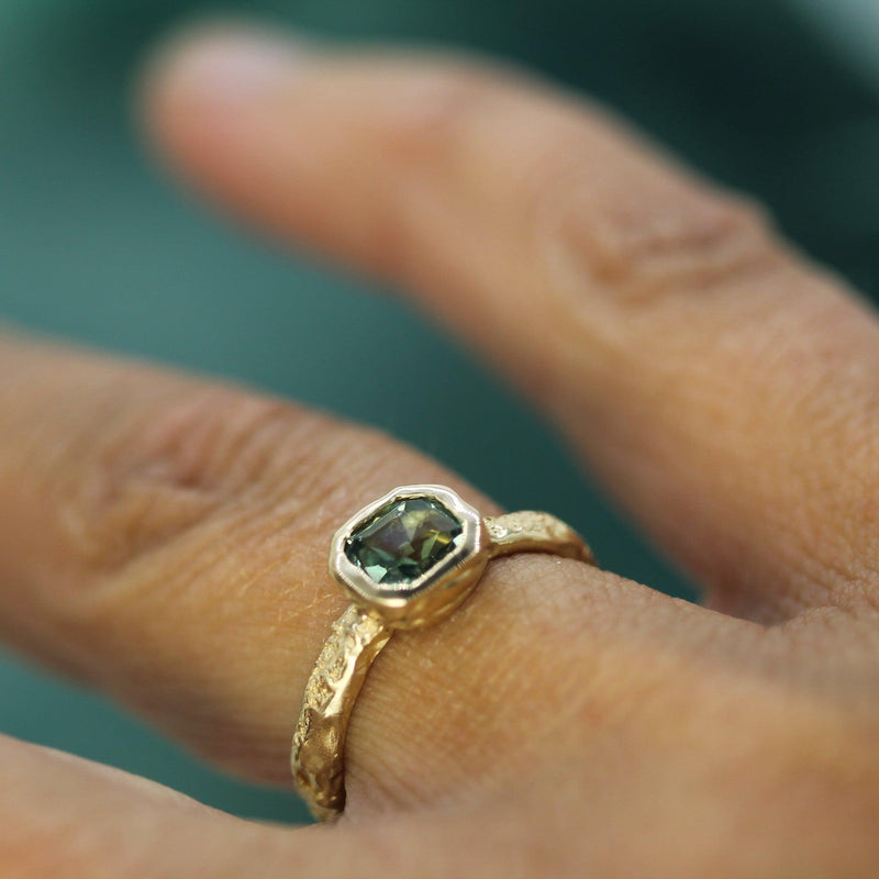 Gold ring with a teal sapphire on a finger against a blurred background