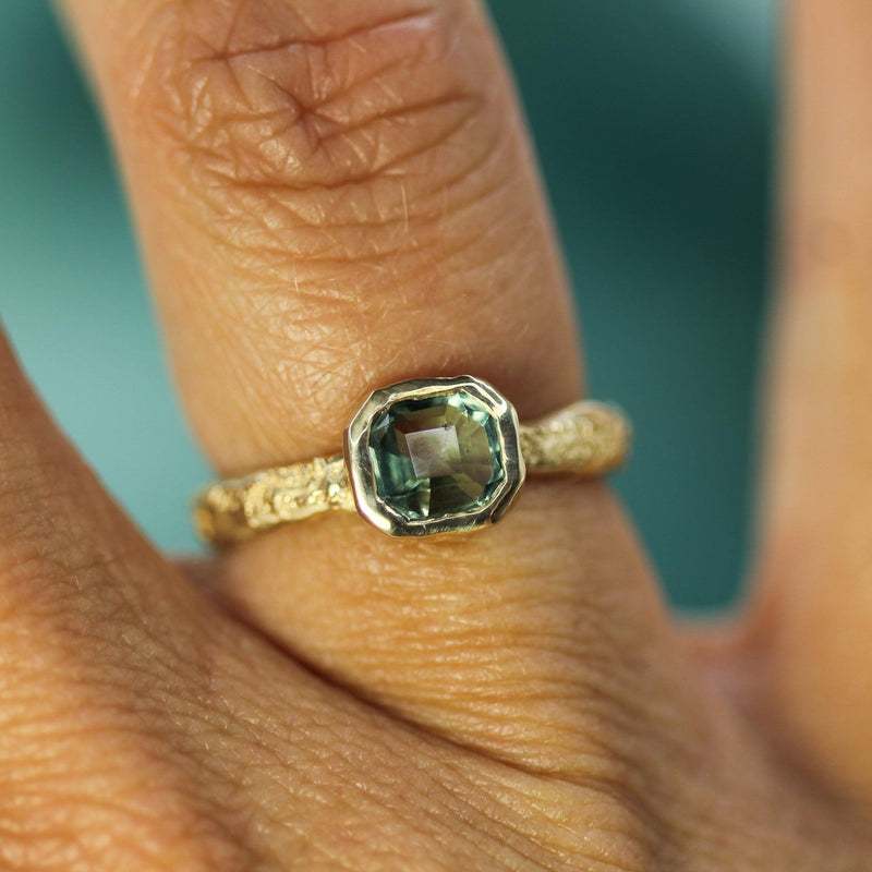 14k yellow gold ring with a teal sapphire  on a finger against a blurred background