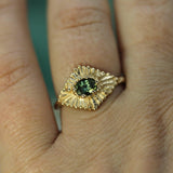 Gold ring with a teal sapphire on a finger against a blurred background