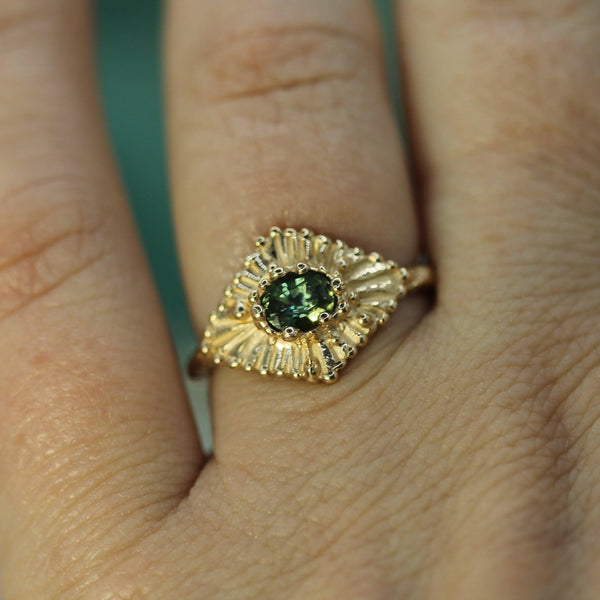 Gold ring with a teal sapphire on a finger against a blurred background