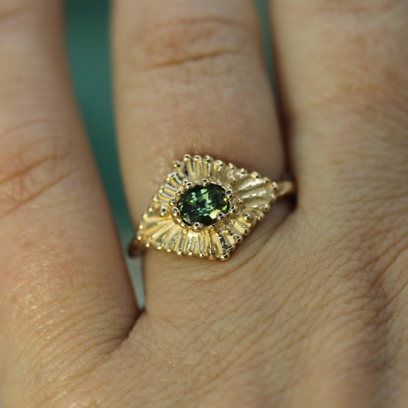 Gold ring with a teal sapphire on a finger against a blurred background
