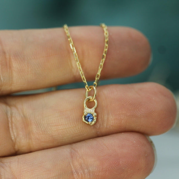 Gold necklace with a blue sapphire pendant held between fingers against a blurred background